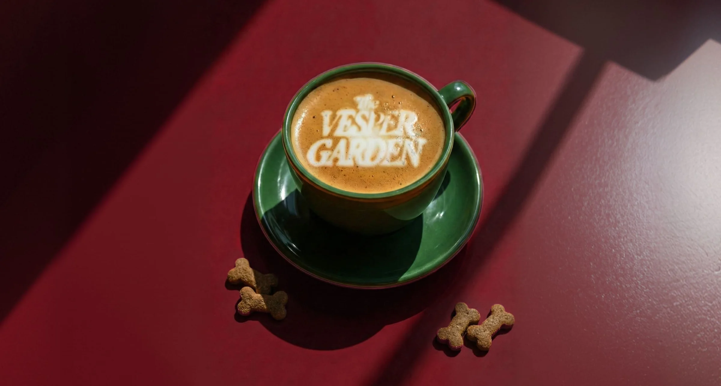 Tasse verte de café avec le logo The Vesper Garden en latte art, biscuits en forme d’os, sur fond bordeaux